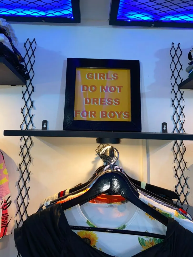 Cadre « Girls do not dress for boys » accroché au-dessus d’un portant de la boutique, sous un néon bleu