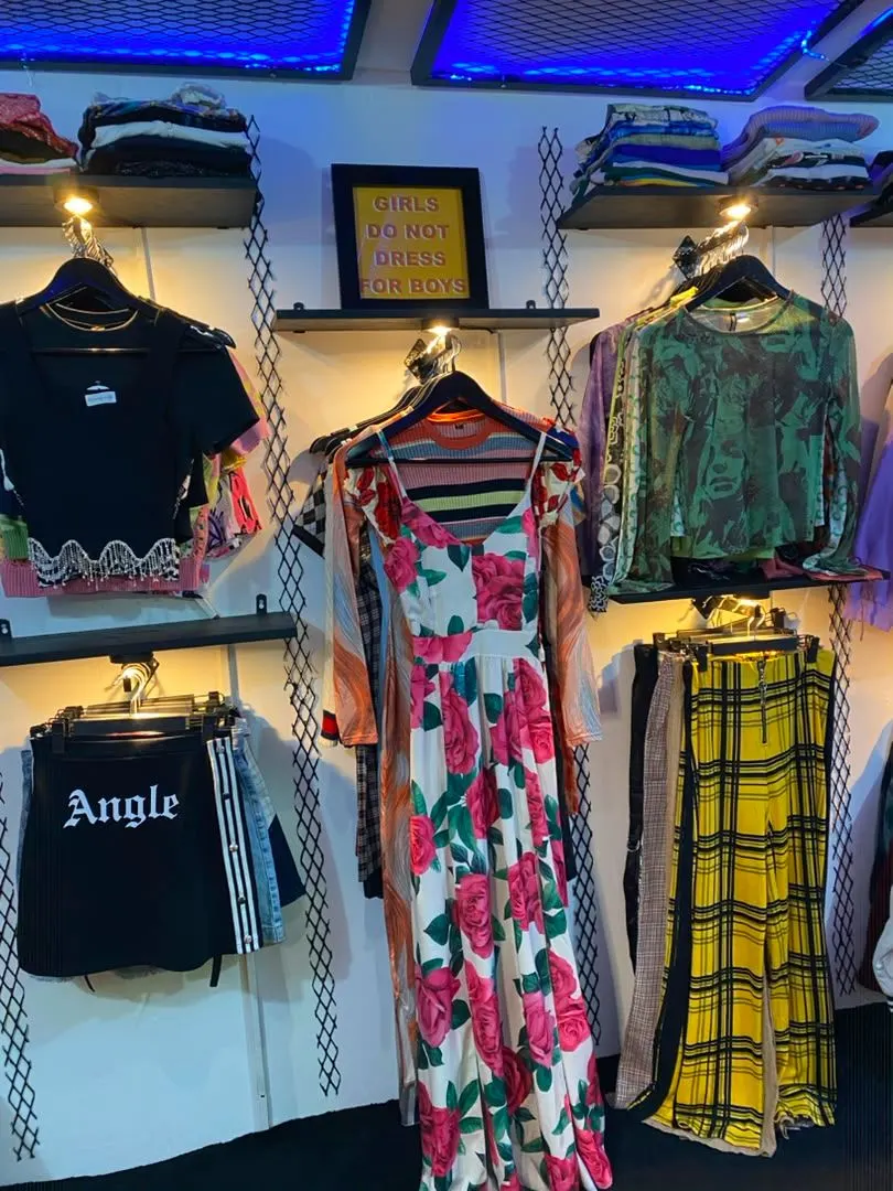 Mur de pièces de friperie : robe à grosses roses, pantalon à carreaux jaune, hauts imprimés et short noir
